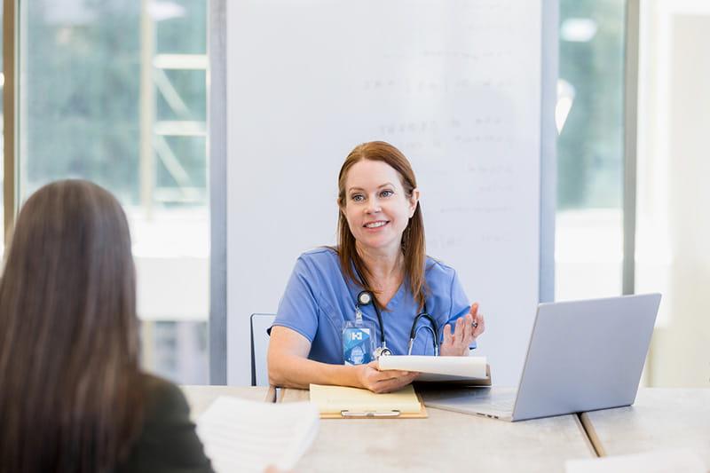 Healthcare professional smiles at colleague while sitting in front of laptop with a clipboard